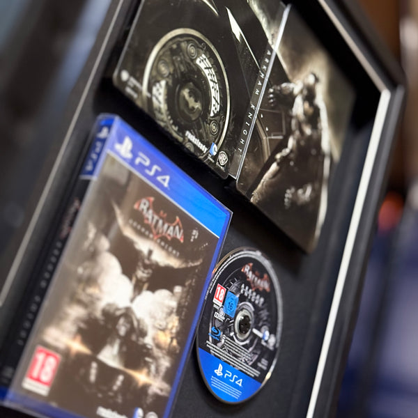 A framed PlayStation 4 game collection display featuring Batman: Arkham Knight game cases, a game disc, and a PS4 console inside a black shadowbox frame.